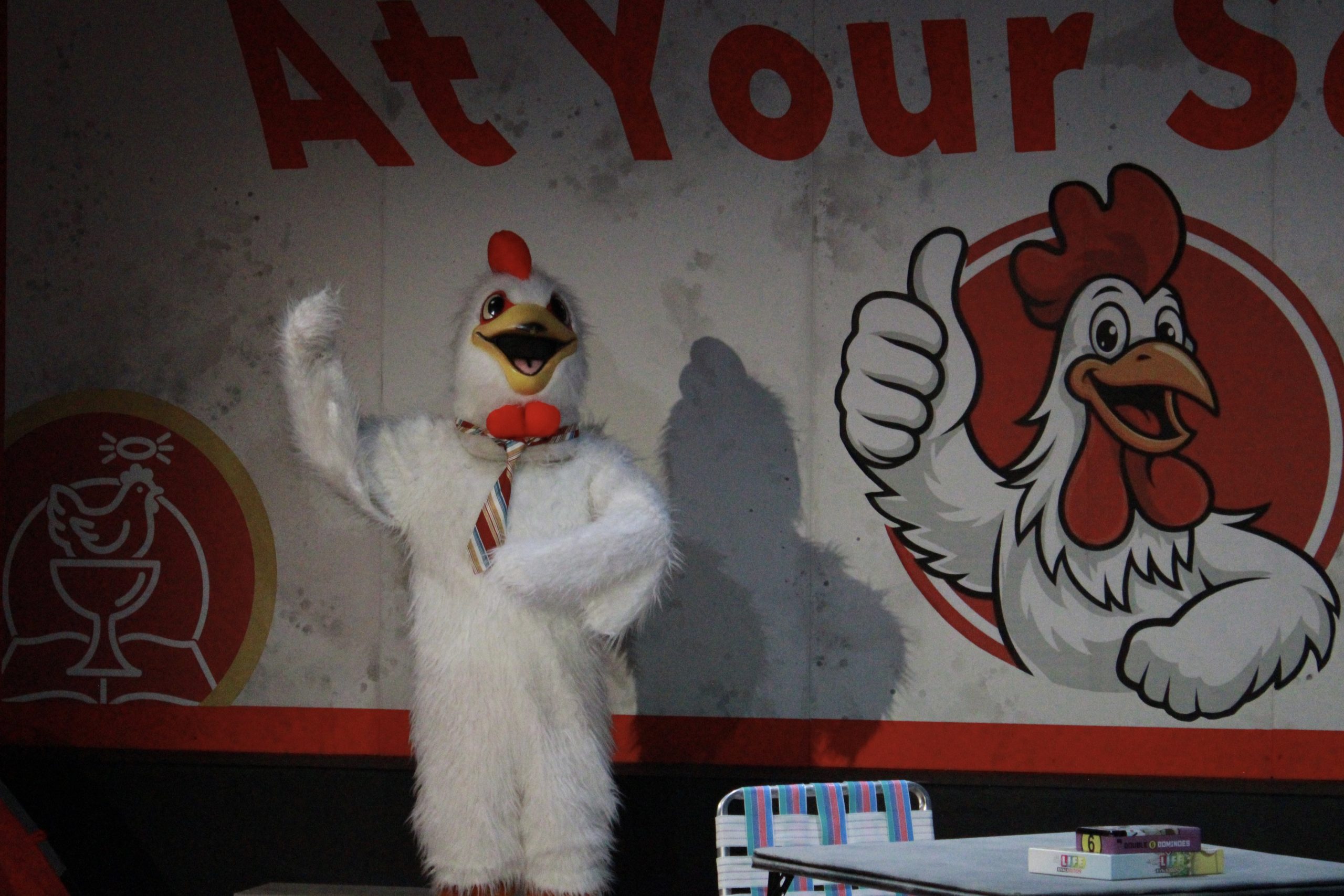 A costume chicken standing with a thumbs up pose, in front of a Billboard that has the text "At your service" above a cartoon chicken that is also holding up a thumbs up.