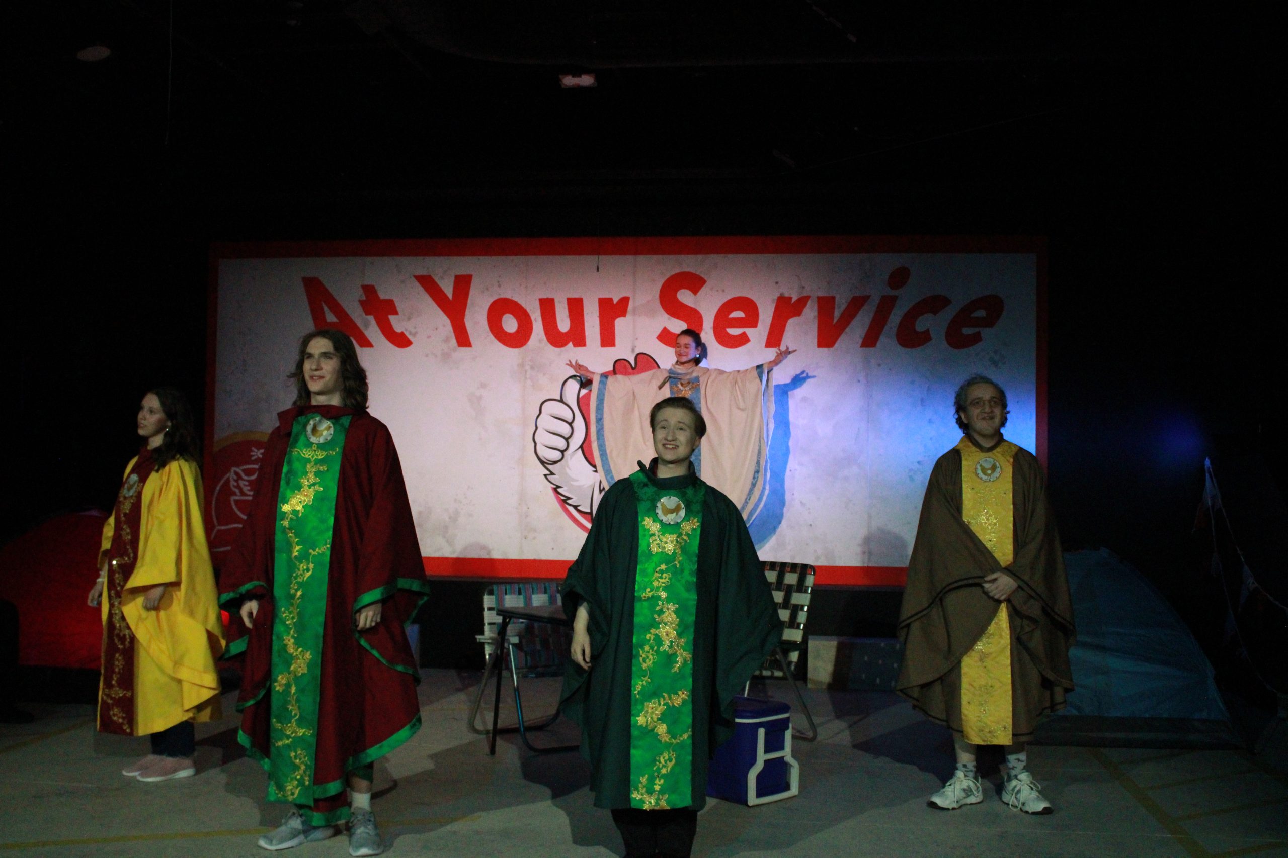 4 actors clad in liturgical robes, standing downstage of a 5th actor in similar robes but in white with blue trim, who has her arms outstretched.